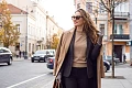 Stilinga ir elegantiška moteris su smėlio spalvos paltu, tamsiu švarku ir akiniais nuo saulės žengia rudeniško miesto senamiesčio gatve