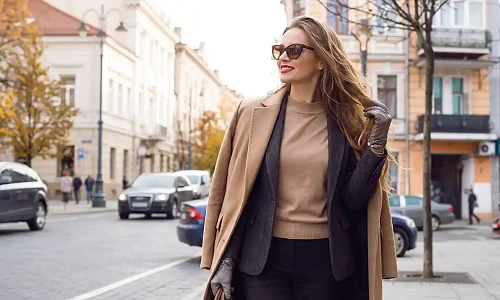 Stilinga ir elegantiška moteris su smėlio spalvos paltu, tamsiu švarku ir akiniais nuo saulės žengia rudeniško miesto senamiesčio gatve